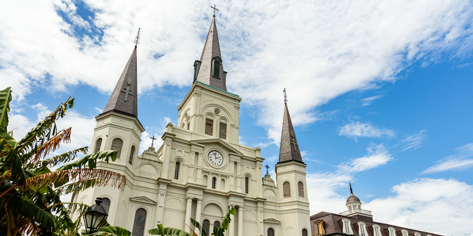 A white cathedral with three spires stands against a blue sky and clouds. 